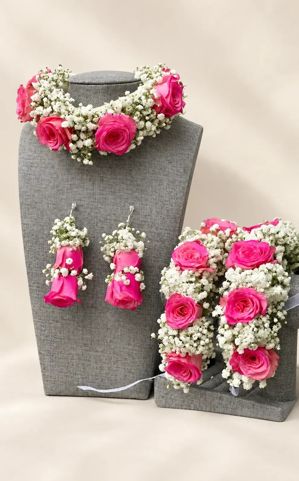 Pink rose and white baby’s breath necklace, dangling earrings and matching hand jewellery displayed on grey stands.
