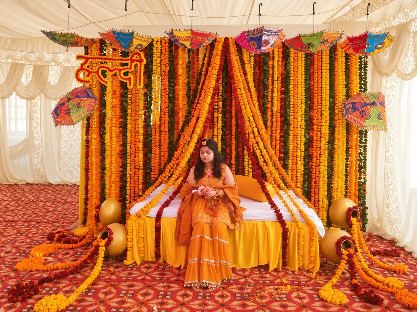 Orange and yellow marigold haldi stage with colourful umbrellas, yellow seating, decorative pots and a red patterned carpet.